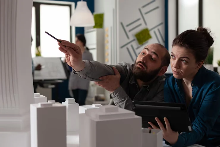 Two property analysts reviewing white building models and tablet while pointing at architectural plans in office