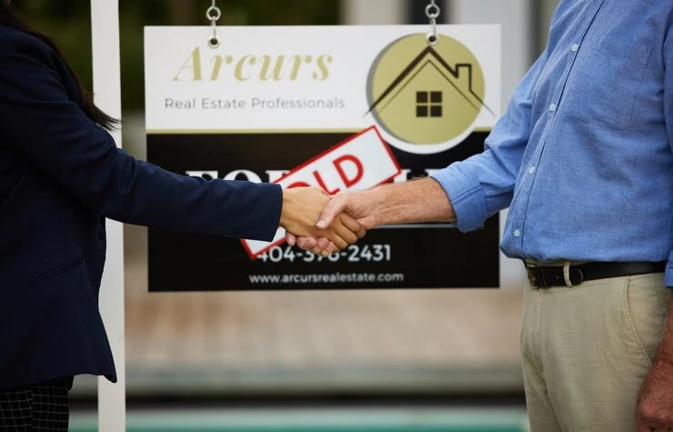 Two professionals shaking hands in front of sold real estate sign completing property transaction