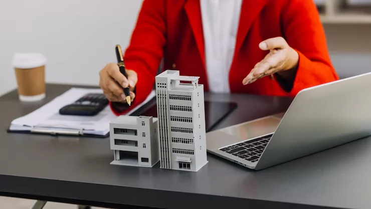 Property analyst in red jacket working with building models, calculator and laptop for investment analysis