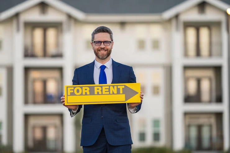Professional in business suit holding yellow "For Rent" sign in front of modern apartment building