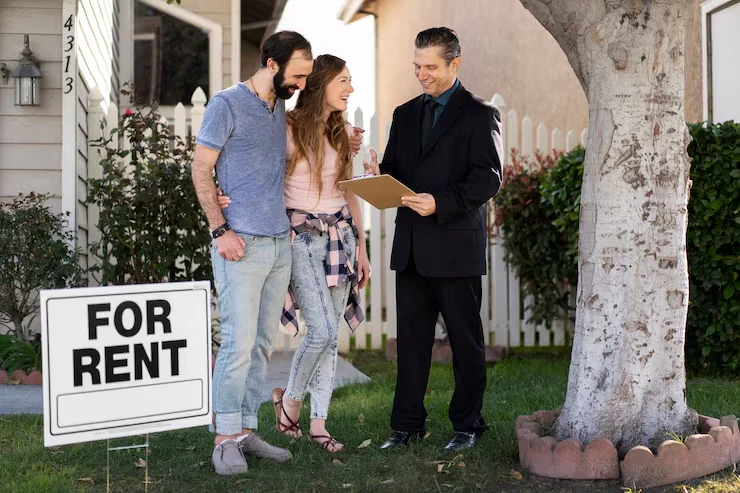Property agent presenting rental agreement documents to young couple standing beside for rent sign