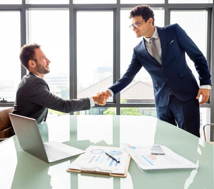 Two businessmen shaking hands over conference table with financial documents in modern office