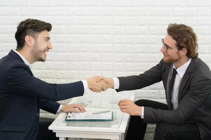 Two businessmen shaking hands over signed property contract on white desk in modern office