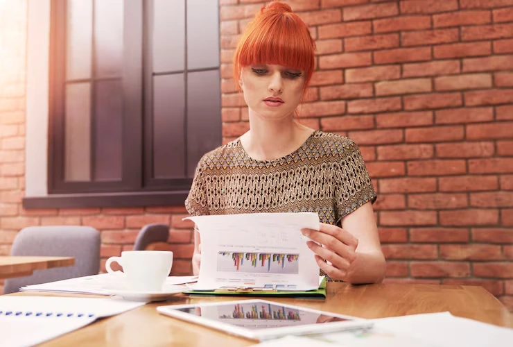 Woman with red hair reviewing property investment charts and data reports at wooden desk with coffee cup