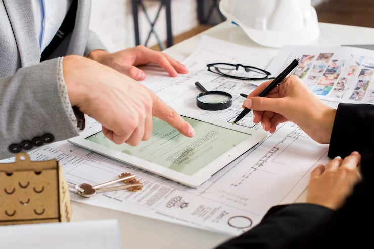 Two professionals analyzing property documents and floor plans on desk with magnifying glass and tablet