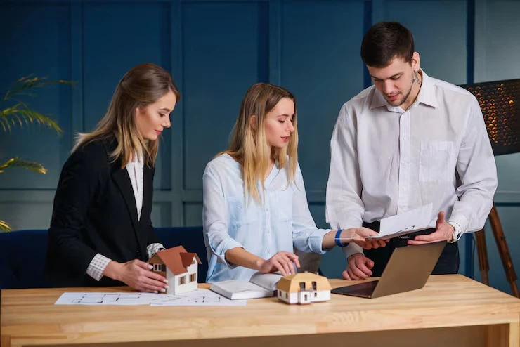 Three property advisors examining house models and reviewing management documents on laptop at conference table