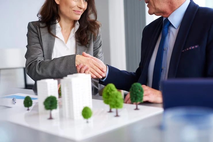 Business professionals shaking hands over architectural model with miniature trees during property deal