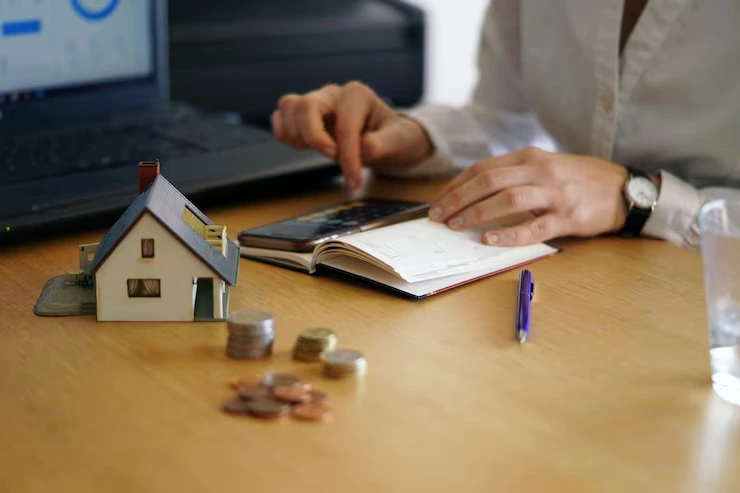 Property manager calculating rental income using calculator with house model, coins, and financial documents on desk