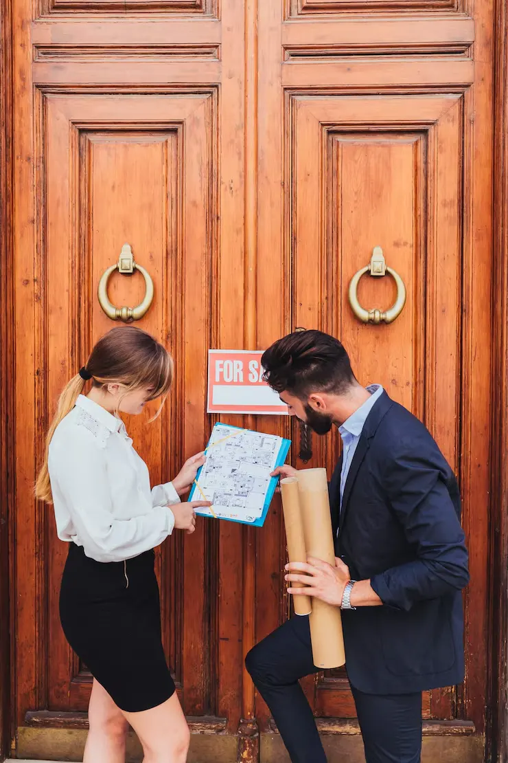 Property agents reviewing floor plans and documents outside building with for sale sign