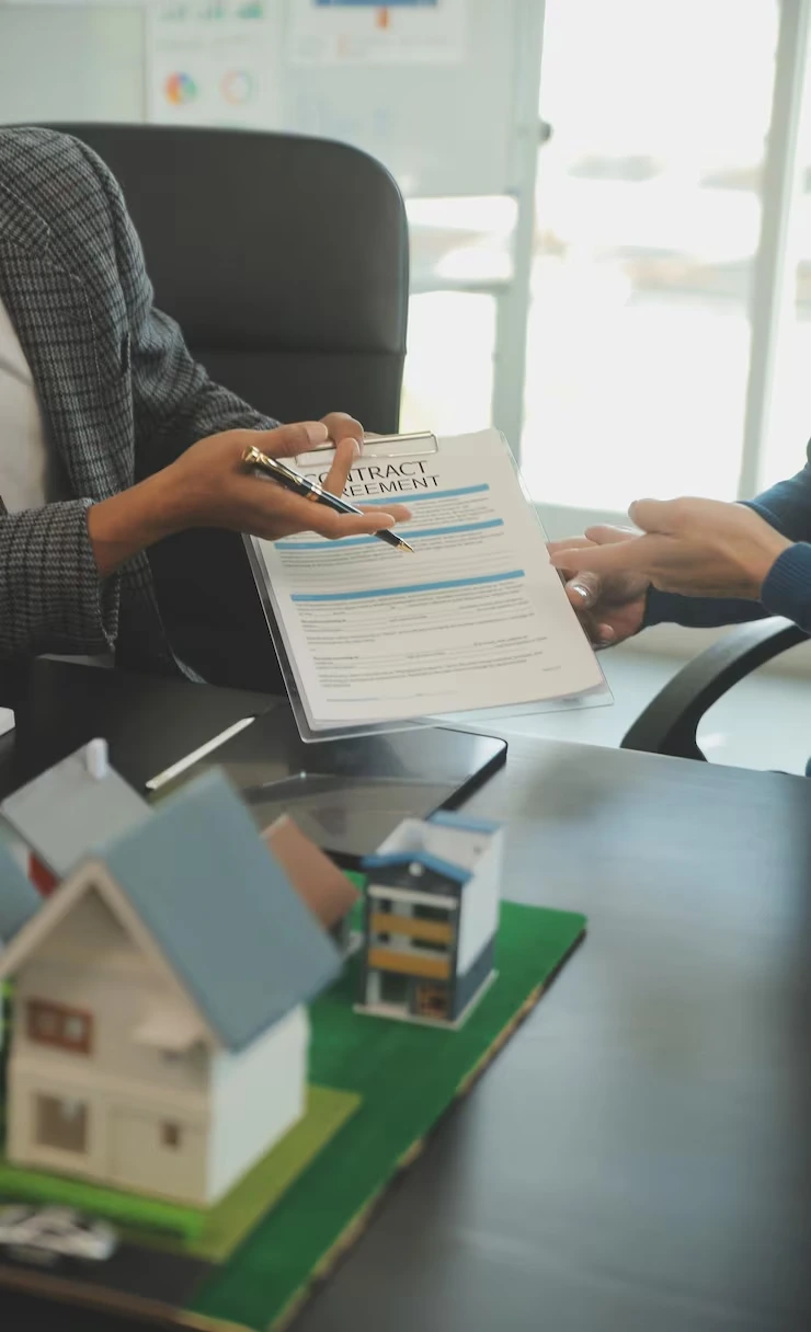 Property agent explaining rental contract agreement to client with house model on office desk