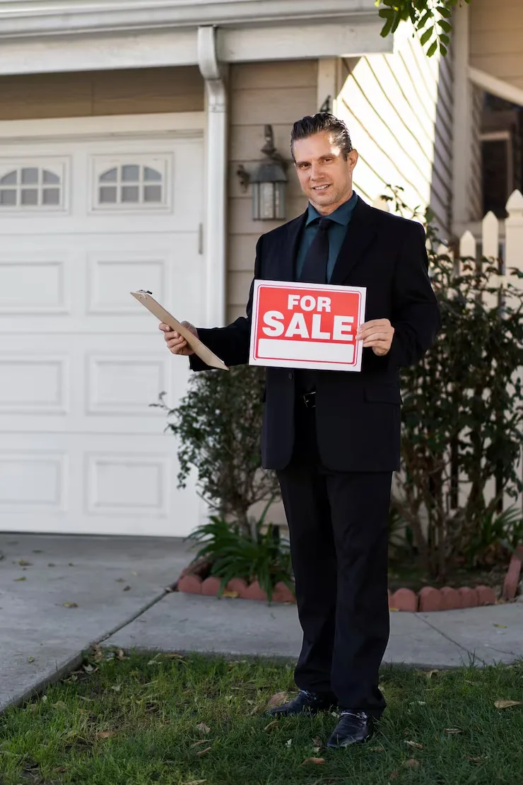 Professional property agent in dark suit holding red for sale sign and clipboard outside residential home